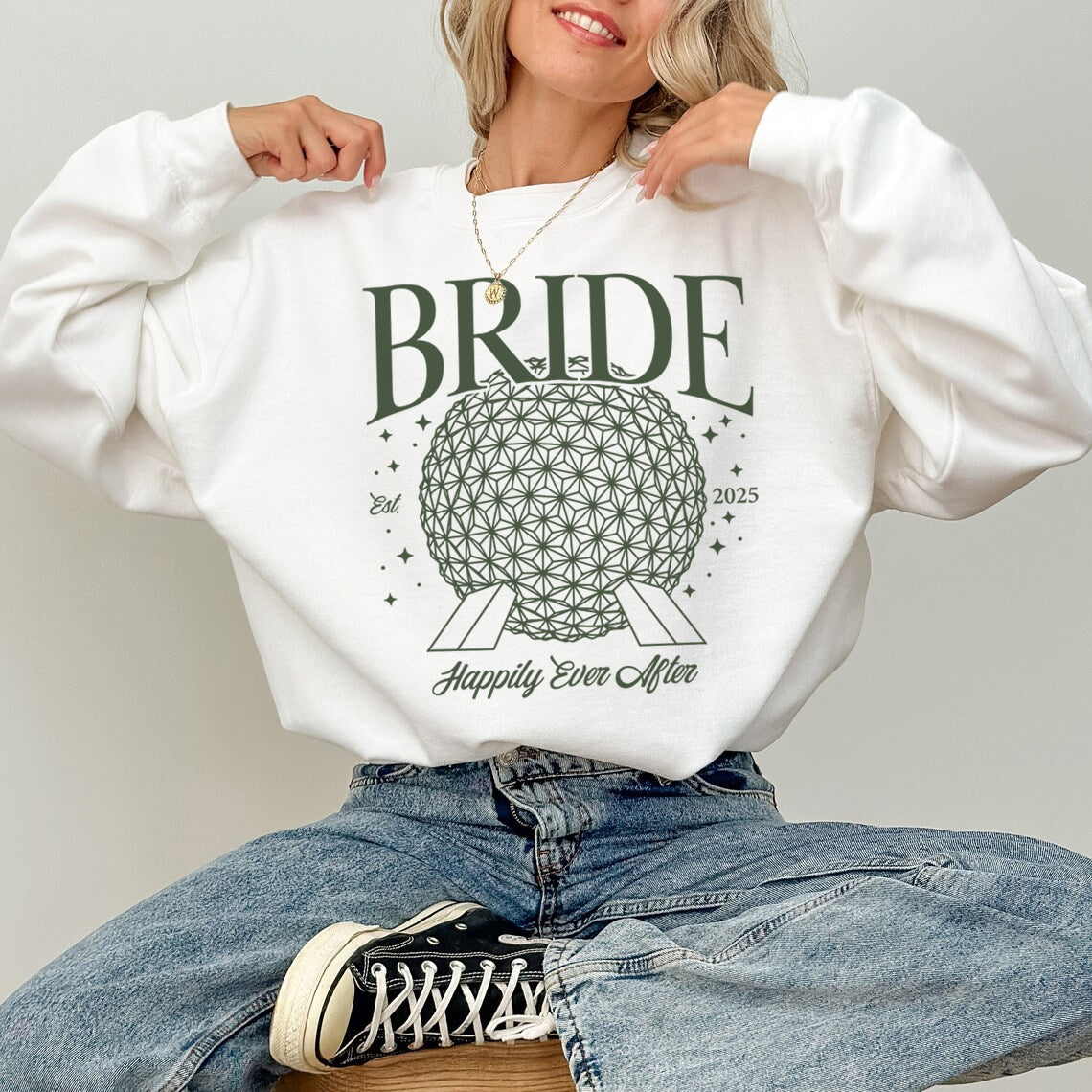 Person wearing a white sweatshirt with 'BRIDE' and wedding-themed design, sitting on a light gray background.